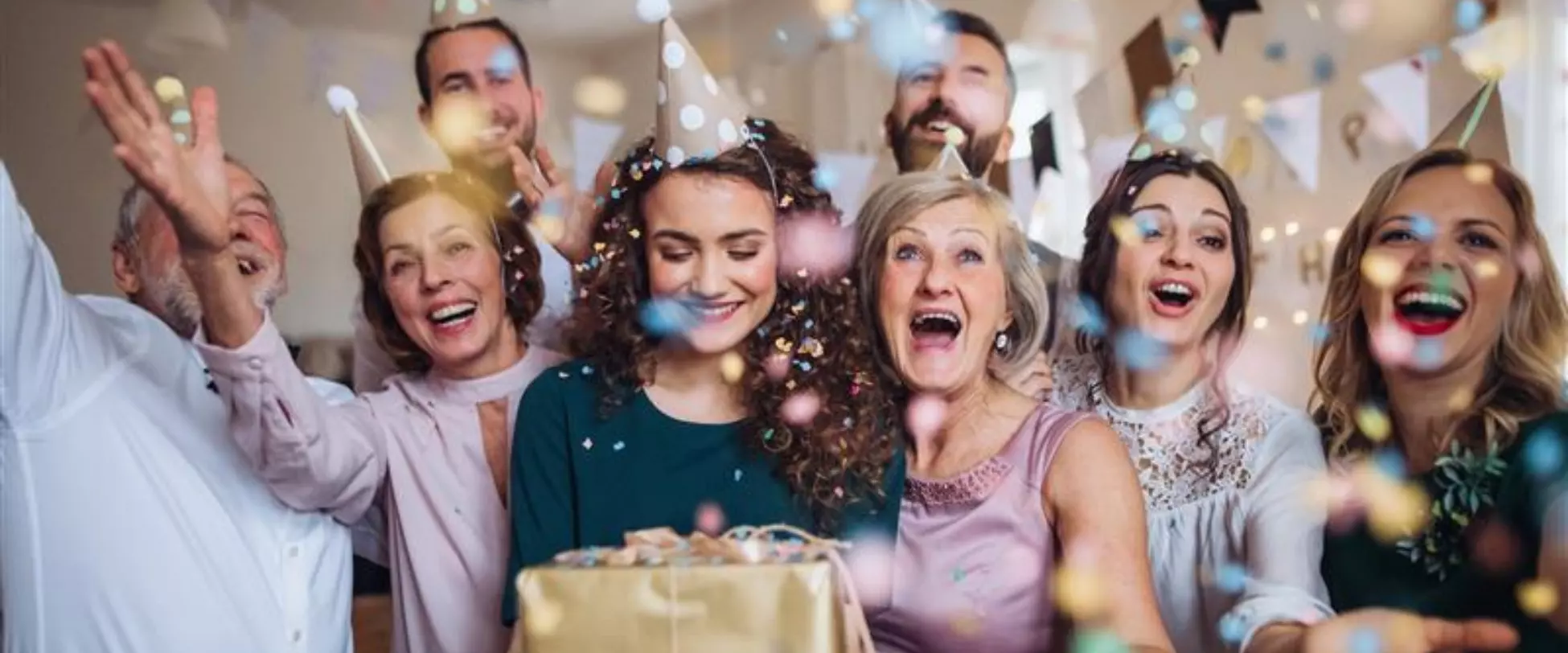 A family are facing the camera and cheering, a woman in the centre is holding a birthday present and a party hat. There is confetti thrown in the air..