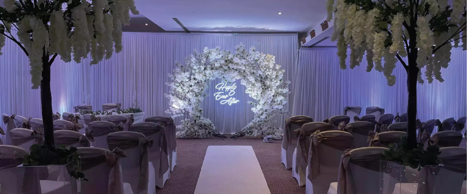 A large room decorated for a wedding. Two trees with white flowers line the aisle alongside chairs set up with white covers and bows. At the from is a white flower arch with the words 