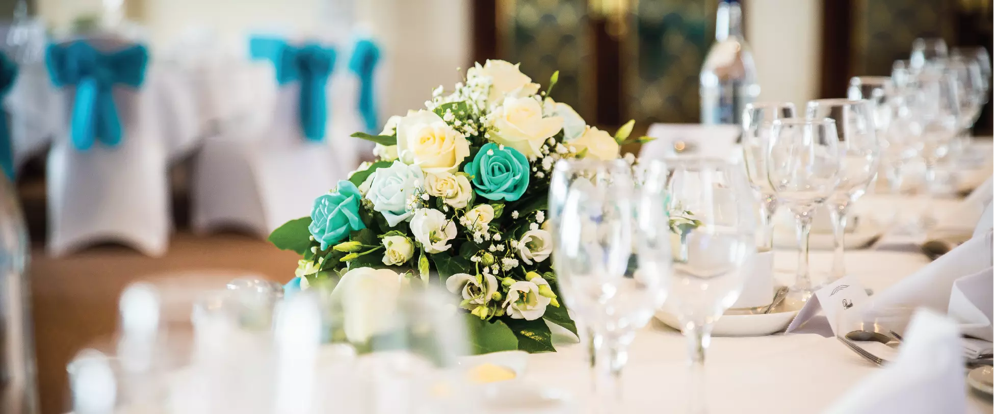 A large room decorated for a wedding. Two trees with white flowers line the aisle alongside chairs set up with white covers and bows. At the from is a white flower arch with the words 